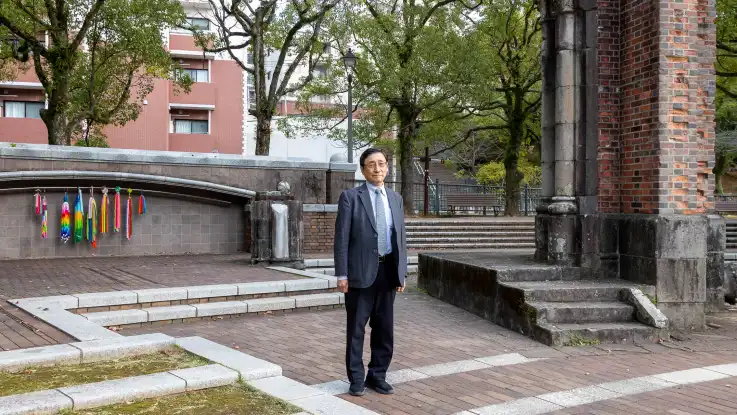 Ein Mann steht neben einer Ruine, hinter ihm hängen bunte Kranich-Girlanden vor einer Steinwand. 