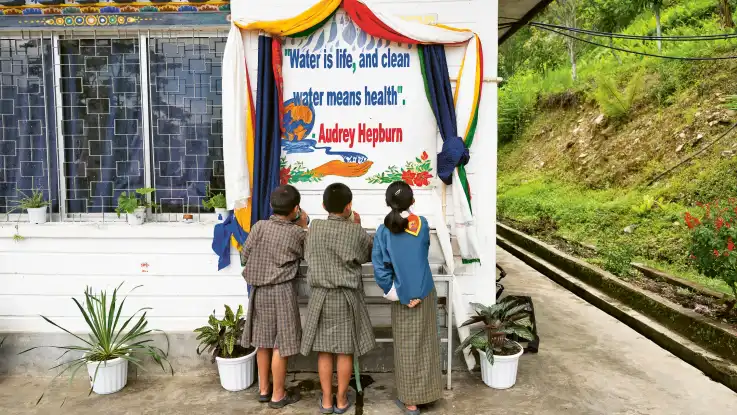 Nun stehen drei trinkende Kinder vor dem Becken und schauen auf ein Plakat mit einem Zitat von Audrey Hepburn.