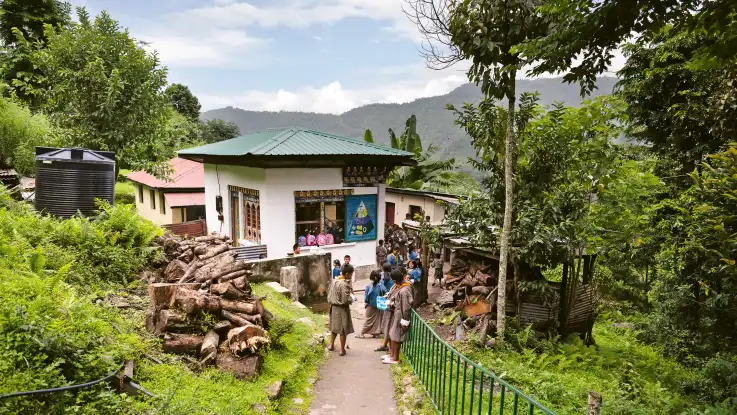 Zwischen kleinen, einstöckigen Häuser stehen zahlreiche Schulkinder. Ein Bergpanorama bildet den Hintergrund. 