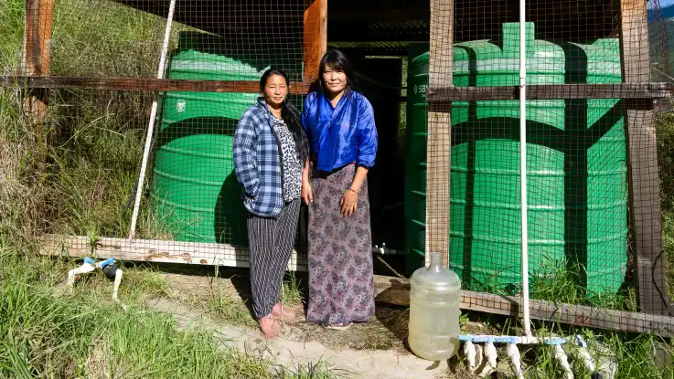 Nun posieren zwei Frauen vor dem Verschlag mit den grünen Wassertanks. 