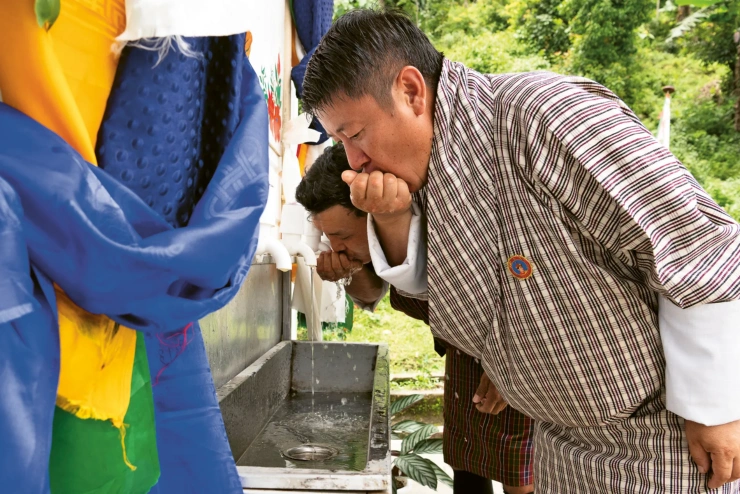 Ein Mann führt mit seiner Hand Wasser aus einem Hahn zum Mund. Hinter ihm beugt sich ein weiterer Mann über das Becken.