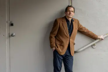 Ein älterer Mann steht mit seinem erwachsenen Sohn vor seinem Gehöft, beide tragen karierte Hemden.