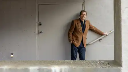 Jon Ippolito steht in einem grau gestrichenen Treppenhaus. Mit der linken Hand hält er den Handlauf, während er nachdenklich in die Kamera schaut.