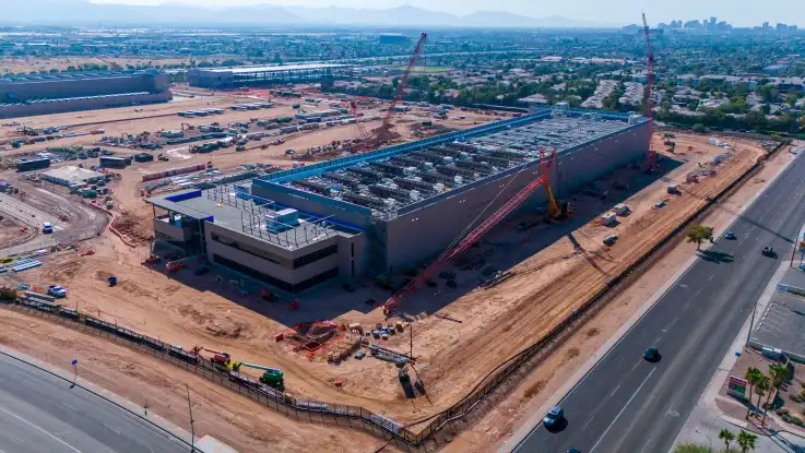 Schräg von oben: ein grau-blaues Gebäude steht umringt von großen Kränen auf rötlich-sandigem Boden.