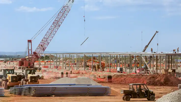 Auf einer belebten Baustelle arbeiten zahlreiche Arbeiter an der Trägerkonstruktion einer weitläufigen Halle. 