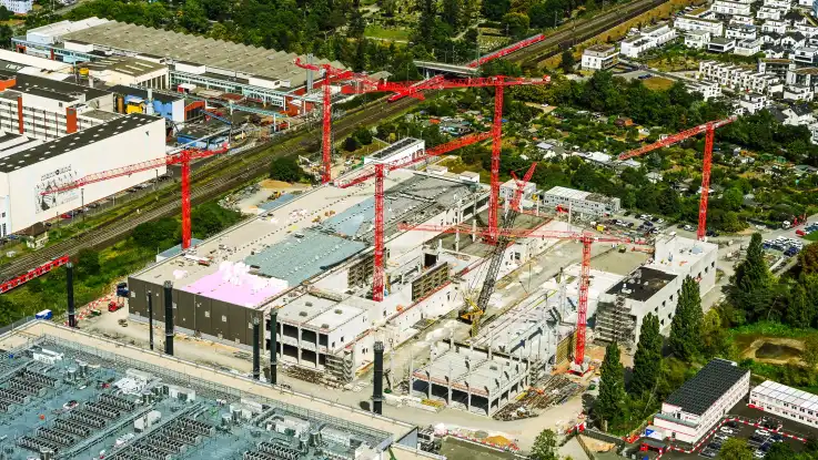 Schräg von oben: Auf einer weitläufigen Baustelle stehen einige Gebäudeteile kurz vor dem Abschluss, während sich andere noch im Rohbau befinden.