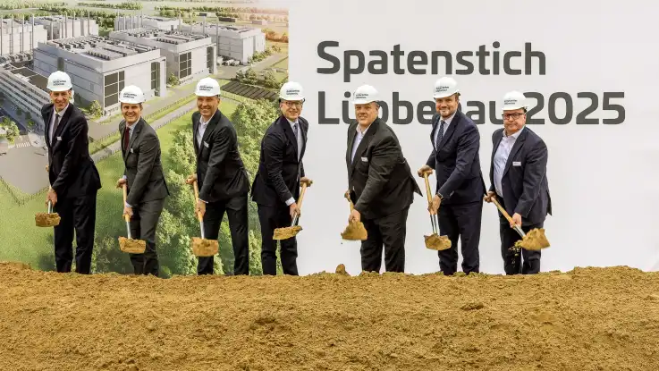 Sieben Männer in Anzügen und mit Bauhelmen auf dem Kopf lächeln angestrengt, während sie synchron mit Spaten Sand ausheben. 