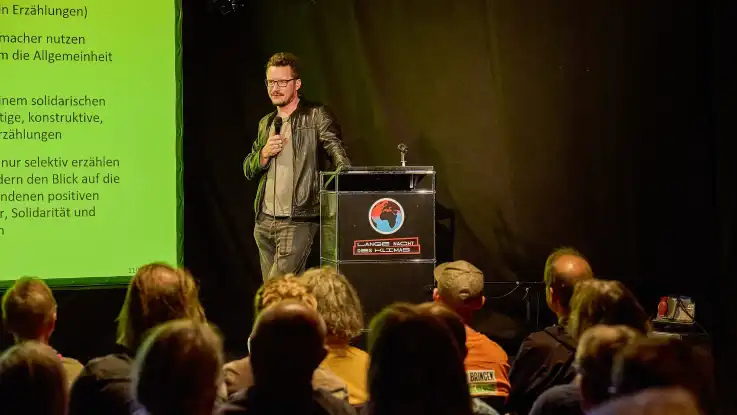 Der Mann lehnt sich locker an das Rednerpult, während er seine Ausführungen fortsetzt. 