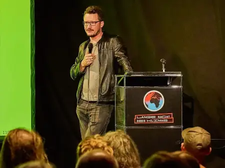 Ein junger Mann steht auf einer Bühne, hält ein Mikro in der Hand und spricht.