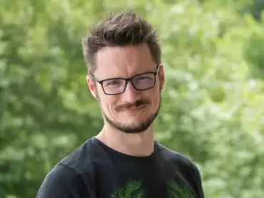 Ein Mann mittleren Alters mit eckiger Brille, Schnauz- und Kinnbart steht im Freien, während er freundlich in die Kamera schaut. 