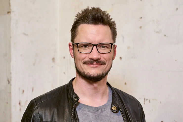 Porträtaufnahme: Ein Mann mittleren Alters mit eckiger Brille, Schnauz- und Kinnbart lächelt in die Kamera. 