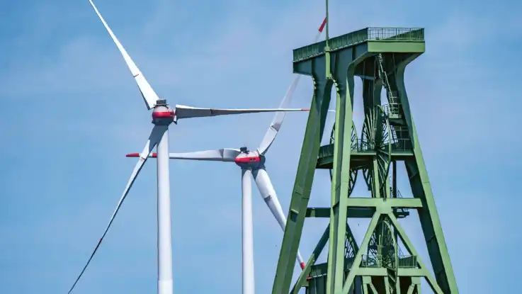 Hinter einem Förderturm aus grün lackiertem Stahl ragen zwei Windkraftanlagen in den Himmel.