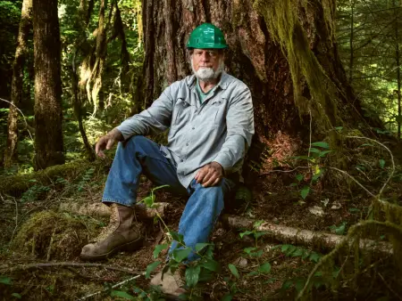 Ein Mann mit grauem Vollbart und grünem Schutzhelm sitzt in Arbeitskleidung an einem Baum gelehnt und schaut in die Kamera.