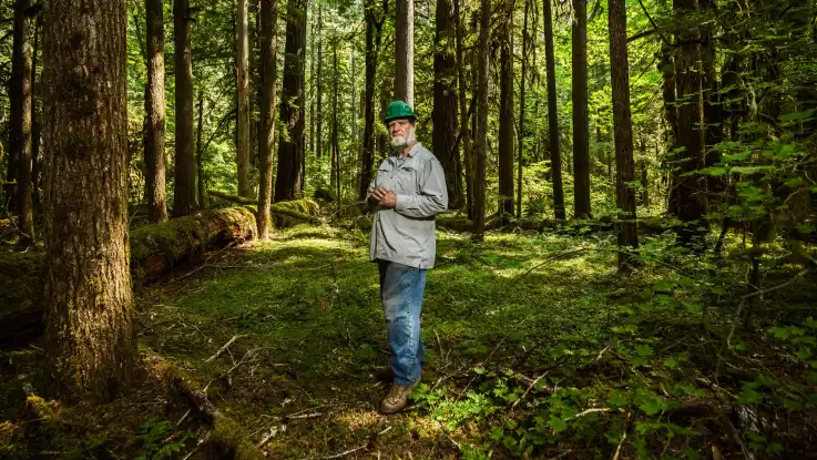 Der Mann mit Vollbart posiert für die Kamera. Sonnenlicht taucht den Wald um ihn herum in ein stimmungsvolles Wechselspiel aus Licht und Schatten. 