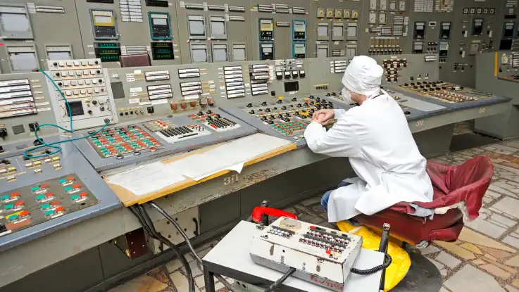 Ein Mann sitzt in weißer Schutzkleidung an einem überdimensionierten Kontrollpult mit Dutzenden Knöpfen. 