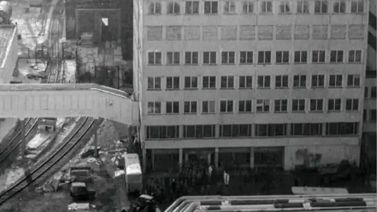 Schwarz-Weiß-Aufnahme: Aus einem mehrstöckigen Haus führt eine Gebäudebrücke über angrenzende Schienen. 