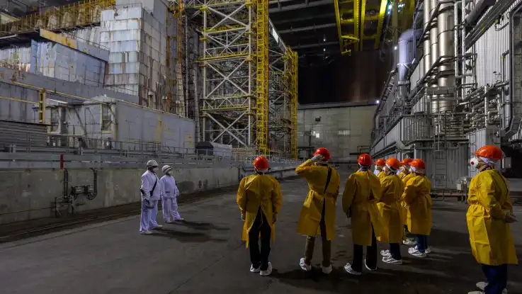 In einer riesigen Halle stehen zwei Männer in weißer Schutzbekleidung etwas abgesetzt von einer Gruppe in gelber Schutzbekleidung. 