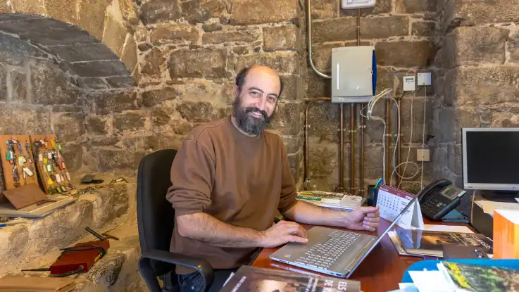 Ein Mann mit Glatze und Vollbart sitzt an einem Schreibtisch vor einer Steinwand und lächelt vollmundig. 