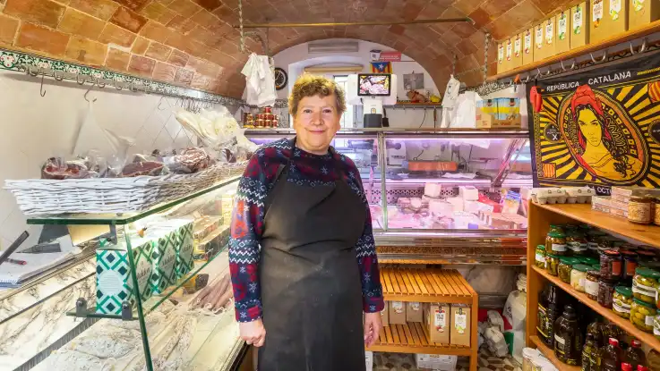 In einem kleinen Laden mit Gewölbedecke steht eine Frau in Schürze und mit kurzem, lockigem Haar vor einer Theke mit Wurst- und Käsespezialitäten. 