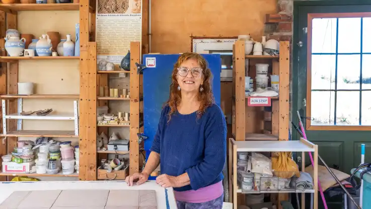 Eine Frau mit langen, roten Haaren steht vor einem blauen Brennofen, drumherum stehen Holzregale mit verschiedenen Töpferwaren. 