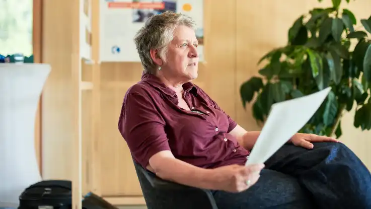  Einzelbild 4: Eine Frau mit Kurzhaar-Frisur hält ein Blatt Papier in der einen Hand, während sie konzentriert nach vorne schaut.