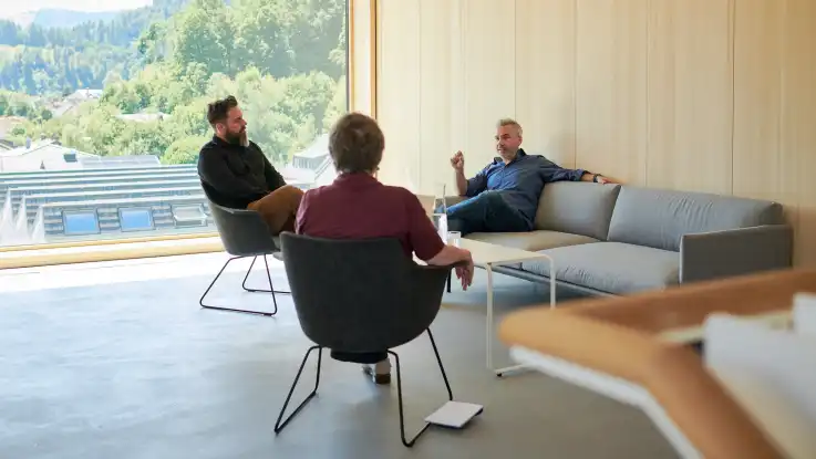 Zwei Männer und eine Frau sitzen entspannt in einem minimalistisch eingerichteten Raum mit vollverglaster Wand. 