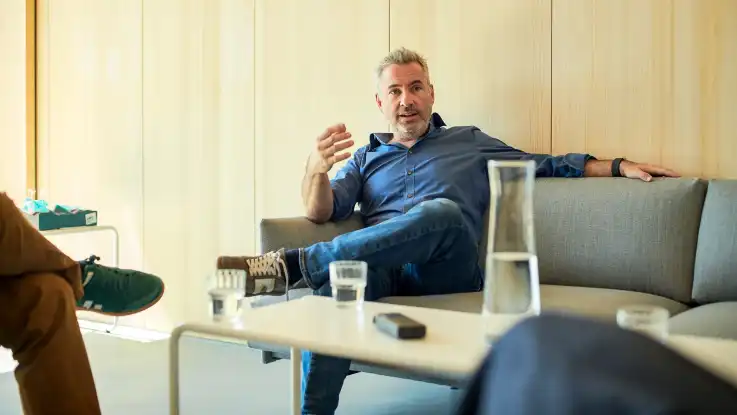 Ein Mann mit grauen Haaren und Drei-Tage-Bart sitzt mit locker übereinander geschlagenen Beinen auf einem grauen Sofa.