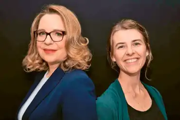 Rücken an Rücken stehen Claudia Kemfert und Christine Langen in einem schwarzen Fotostudio und schauen in die Kamera. 