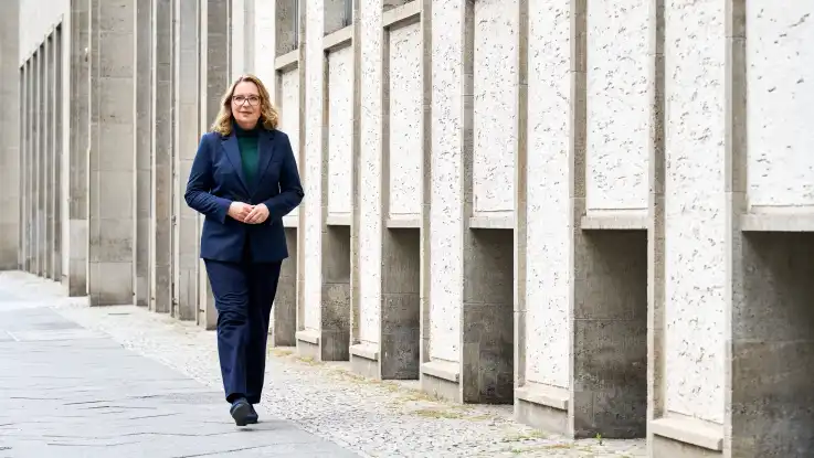 Entlang einer grauen, rhytmisch eintönigen Gebäudeflucht läuft eine Frau mit blonden Haaren, Brille und in einem eleganten Anzug.
