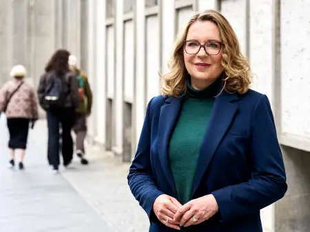 Frau Kemfert steht auf der Straße, an einer grauen Gebäudeflucht. Im Hintergrund geht eine Gruppe von Passanten.