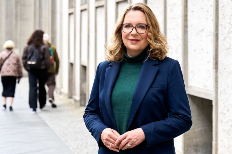 Frau Kemfert steht auf der Straße, an einer grauen Gebäudeflucht. Im Hintergrund geht eine Gruppe von Passanten.