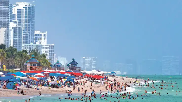 Vor einem Hochhauspanorama liegen unzählige Menschen dicht gedrängt zwischen Sonnenschirmen an einem Sandstrand mit türkisfarbenem Wasser.