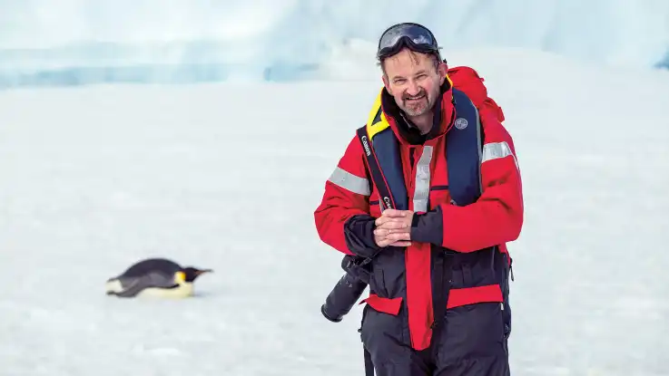 Ein Mann in roter Schneekleidung schaut mit zusammen gekniffenen Augen in die Kamera, während hinter ihm ein Pinguin auf dem Bauch liegt.
