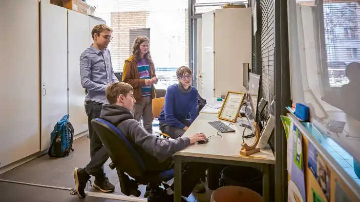 Vier Jugendliche in einem Klassenraum sitzen und stehen an einem Tisch mit Computer und Dokumenten und führen eine Besprechung durch.