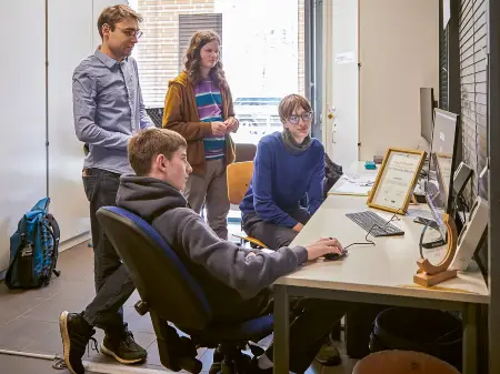 Vier Jugendliche in einem Klassenraum sitzen und stehen an einem Tisch mit Computer und Dokumenten und führen eine Besprechung durch.