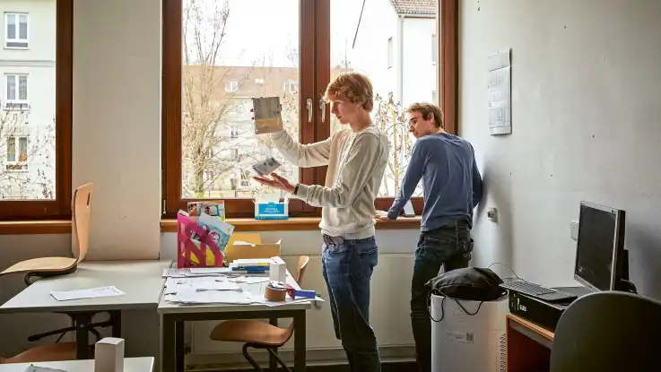 Vor den Heizkörpern unter den großen fesntern des Klassenzimmers arbeiten zwei junge Männer konzentriert an der Installation von neuen Heizungs-Thermostaten.