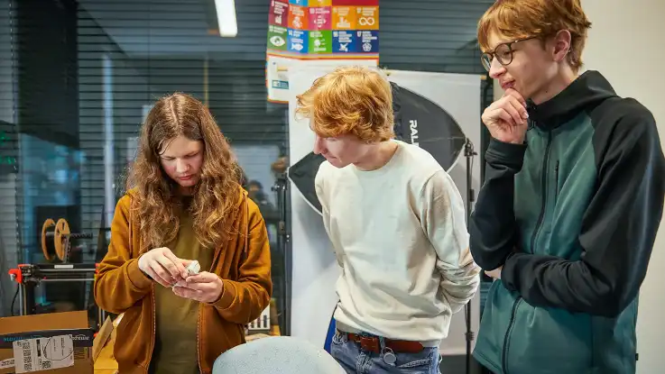 Drei Jugendliche stehen in einem Büro, ein Jugendlicher hält einen kleinen Gegenstand in den Händen, die anderen beobachten aufmerksam.