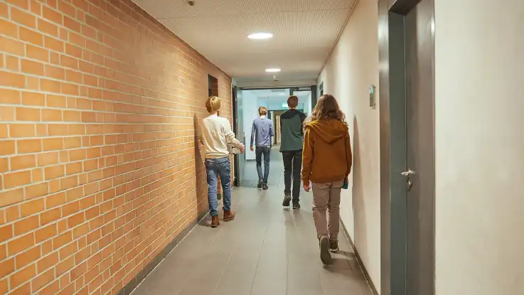 Ein Flur in einer Schule mit vier Jugendlichen, die sich von der Kamera weg in Richtung eines hellen Ausgangs bewegen. 