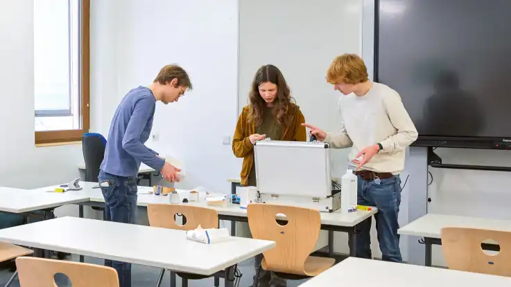 In einem hellen Klassenzimmer stehen drei Junge Männer an einem Tisch und haben allerhand Utensilien aus einem Koffer auf den Tisch ausgebreitet.