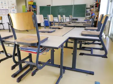 Ein leerer Klassenraum mit mehreren aneinandergereihten Tischen und darauf gestellten Holzstühlen mit Metallgestell.