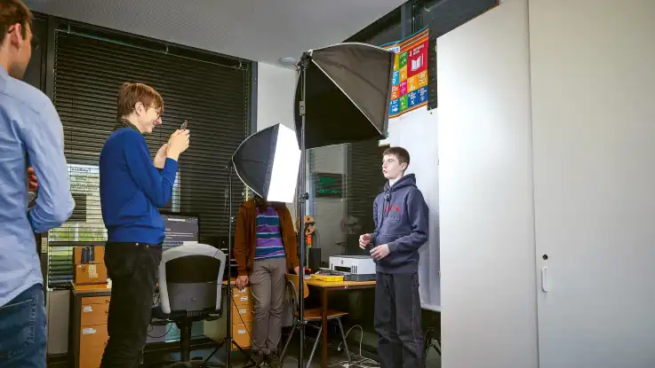 Mehrere Jugendliche stehen in einem Klassenraum, einer wird vor Studiolicht und Softbox fotografiert, während andere zusehen.