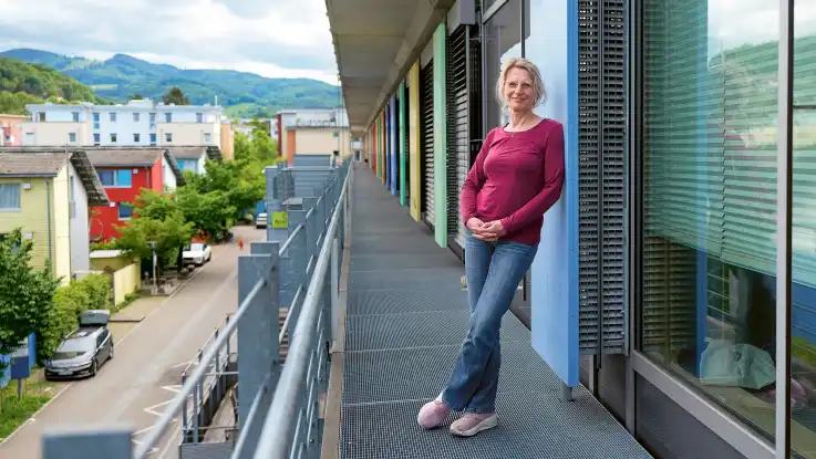 Auf einem Balkon steht eine Frau locker angelehnt an die Fassade und schaut offen in die Kamera.