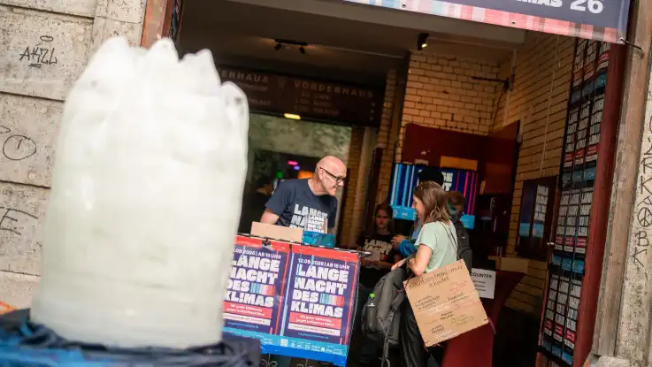 Mann mit Brille und schwarzem T-Shirt steht hinter einem Tisch mit Plakaten vor einem Eingang, eine Frau mit Rucksack und Papiertüte spricht mit ihm. Links im Vordergrund steht eine Eisskulptur.