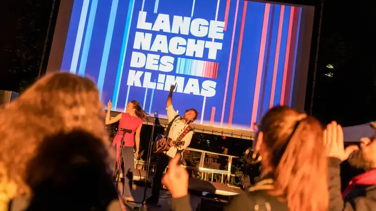 Musiker auf Bühne vor großem Bildschirm mit Schriftzug «Lange Nacht des Klimas» und Publikum im Vordergrund.
