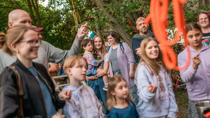 Gruppe von Erwachsenen und Kindern im Freien vor Bäumen, einige lächeln und schauen in Richtung Kamera.