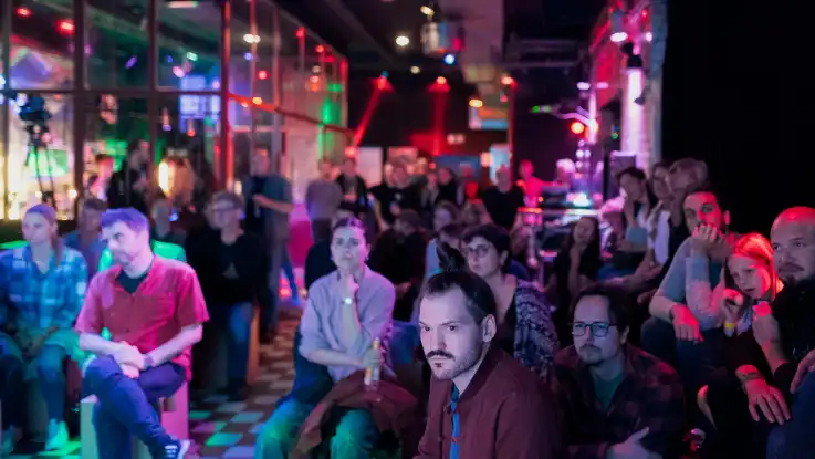 Gruppe von Menschen sitzt in einem dunklen Raum mit bunter Beleuchtung und schaut nach vorne.
