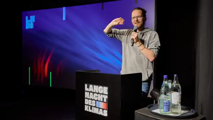 Mann mit Brille und grauem Pullover hält Mikrofon und steht an einem Rednerpult mit der Aufschrift «Lange Nacht des Klimas» vor einem dunkelblauen Hintergrund mit farbigen Lichtstreifen.