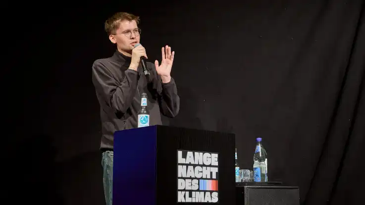 Mann mit Brille und Mikrofon steht hinter einem schwarzen Rednerpult mit der Aufschrift «Lange Nacht des Klimas» und spricht vor schwarzem Hintergrund.