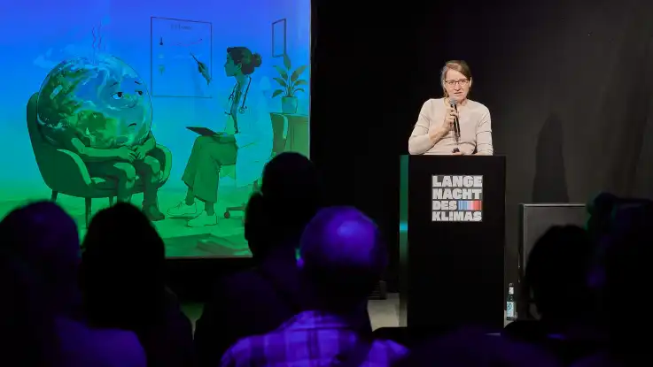 Frau mit Mikrofon steht am Rednerpult mit Aufschrift «Lange Nacht des Klimas» vor Publikum und neben einer Leinwand mit einer Illustration einer Person, die wie ein Erdball aussieht, und einer Ärztin in einem Behandlungszimmer.