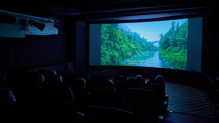 Dunkler Kinosaal mit Zuschauern, die einen Film mit einer Landschaft aus bewaldeten Hügeln und einem Fluss sehen.